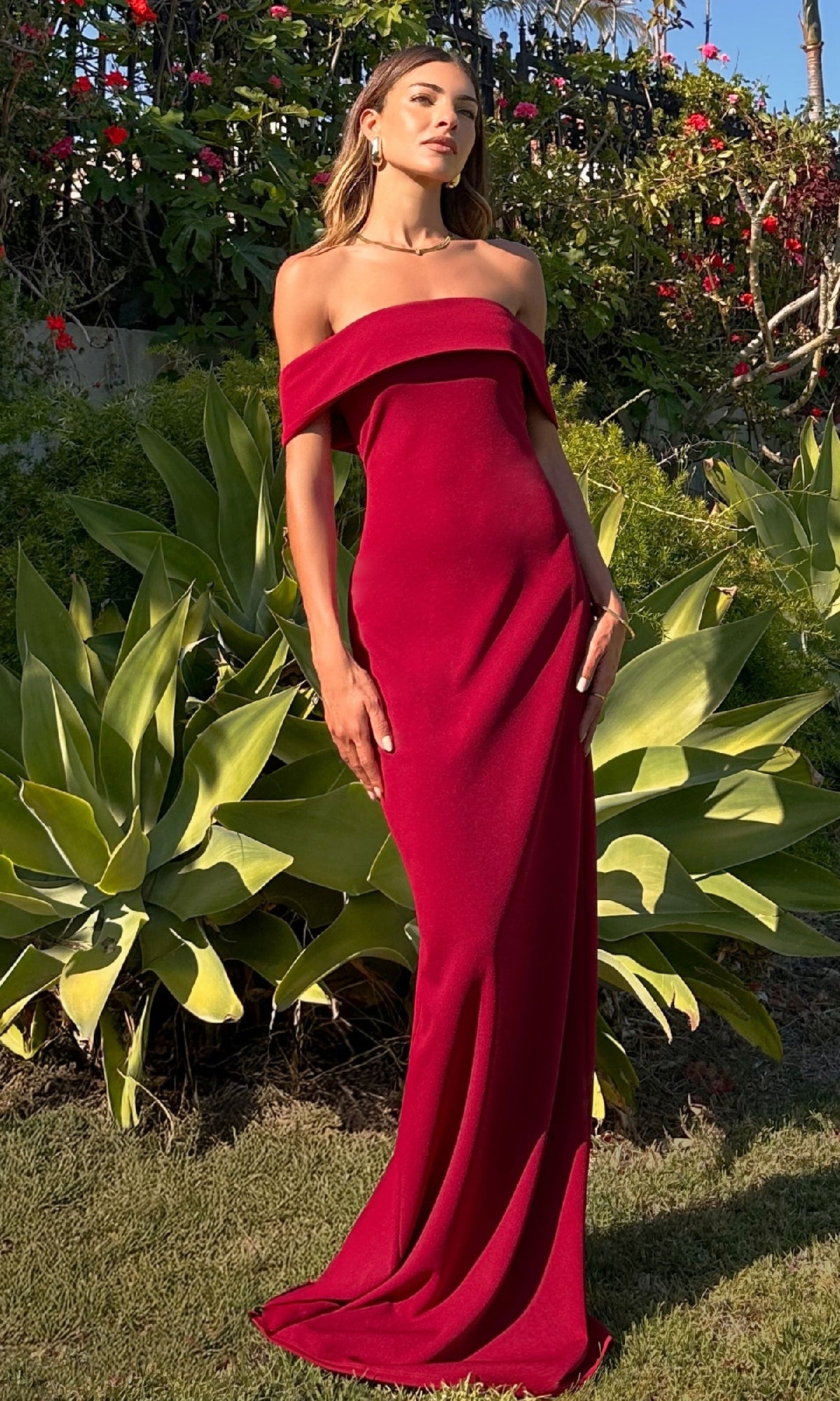 A woman stands outdoors among green plants and flowers, her formal look highlighted by the elegant Off-the-Shoulder Long Prom Dress: Astee T2011 in classic red.